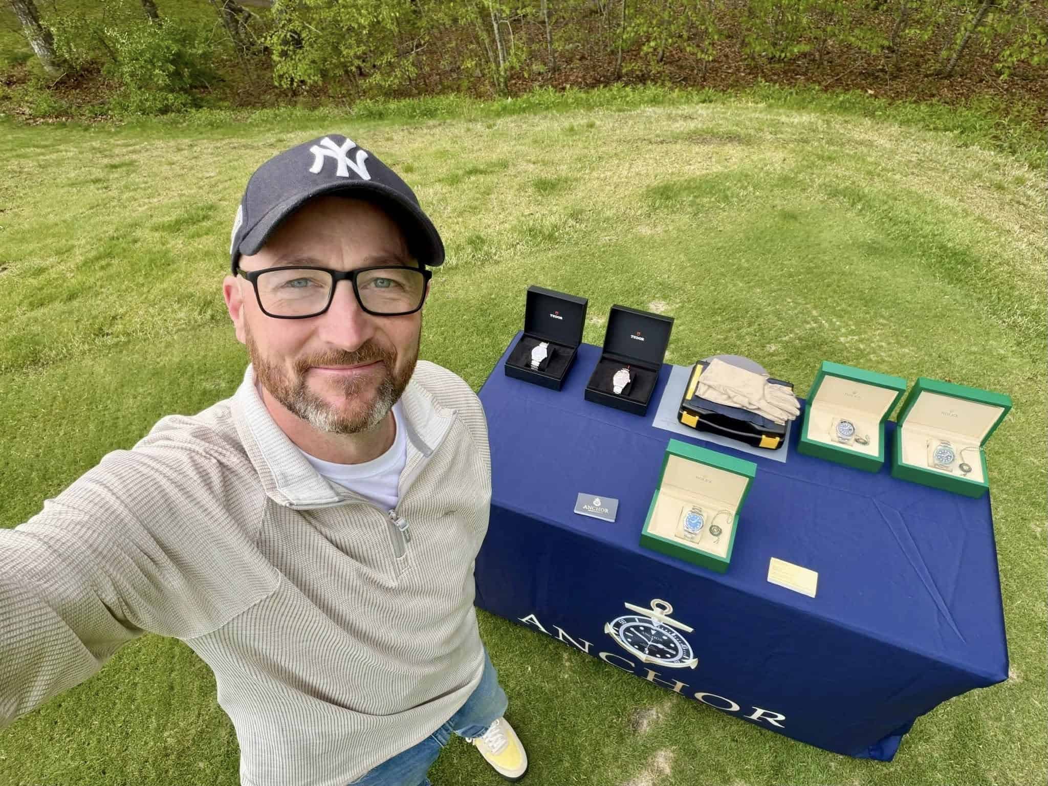 Man in a cap takes a selfie on grass next to a table displaying boxed watches and items with an Anchor logo tablecloth. Trees are visible in the background.